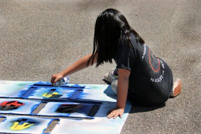 child wearing black t-shirt uses spray paint and cardboard stencil to paint hopscotch squares on pavement