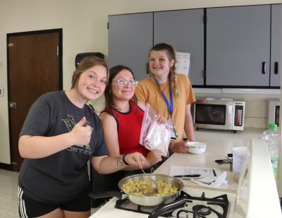 three smiling teenage girls stand beside a stove with a pan full of potatoes, demonstrating their cooking skills. One gives a thumbs up sign and another holds up a packet of recipes.