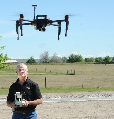 man wearing glasses and black polo shirt pilots a drone in a dirt parking lot near a fenced field