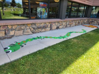 sidewalk painted with green caterpillar with the alphabet written on its body and a colorful butterfly. The sidewalk is bordered by a building with an open sign in the window and green grass