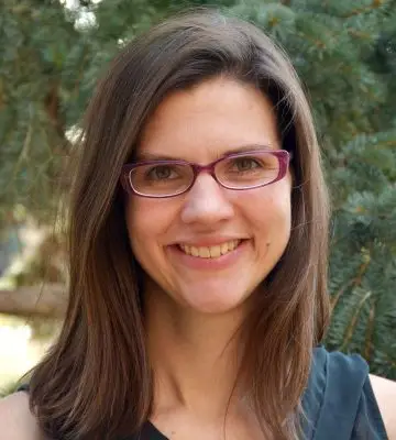 smiling woman with dark hair and purple rimmed glasses stands in front of pine tree