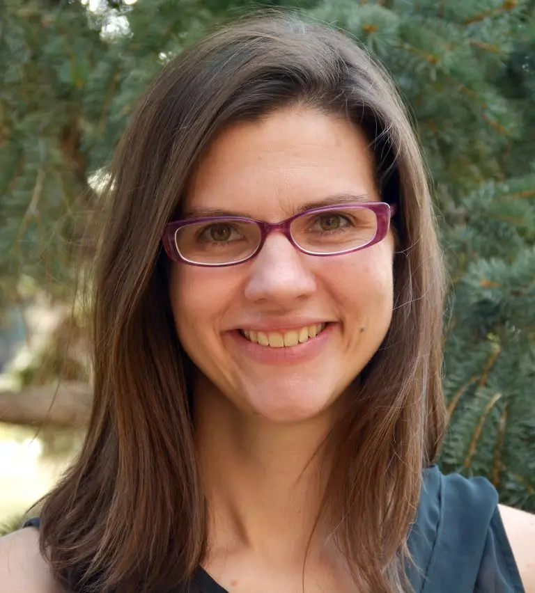 smiling woman with dark hair and purple rimmed glasses stands in front of pine tree