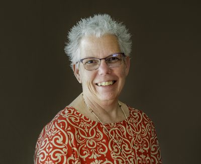 women with short spiky gray hair and glasses smiles for a photo. She is wearing a red floral shirt.