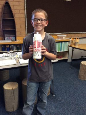 smiling elementary school boy wearing black-rimmed glasses and jeans holds up a white bottle with red lettering and his name, David, in black letters