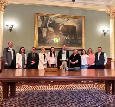ten smiling people stand in a line with Wyoming's governor in the center holding a green sheet of paper. There is a large painting of cows in a gold frame hanging on the wall behind them and a low wooden table in front of them.