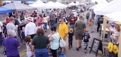 A crowded farmers market on a downtown street with throngs of people moving between two aisles of small, tent-covered booths.