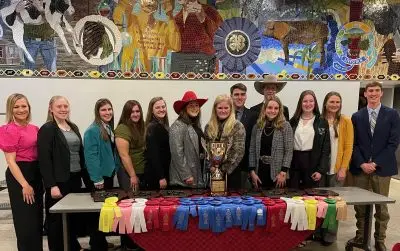 thirteen smiling people stand below a colorful tile mural in front of a plastic table piled with award plaques, ribbons, and trophy cup.