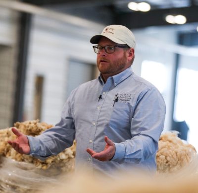man wearing baseball cap, glasses, and blue UW Extension collared shirt delivers a presentation in front of bags of wool