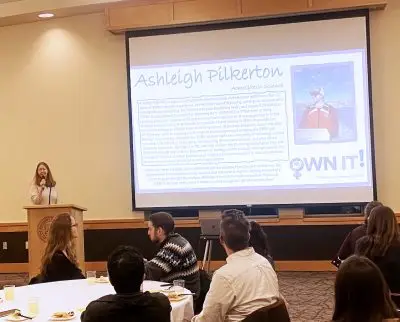 Smiling woman stands at a podium holding a microphone beside a projected Powerpoint presentation. She is facing group of people clustered around a circular table with white tablecloth and plates of food.