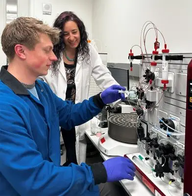 man wearing blue lab coat and gloves inserts a test tube into a protein-purifying machine while a smiling woman in a white lab coat looks on