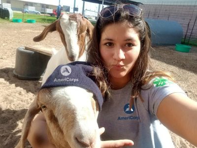 Young woman with dark hair and aviator sunglasses perched on her head and wearing a gray T-shirt with 4-H and AmeriCorps logos takes a selfie with two goats. She. has her arm around one wearing an AmeriCorps knit cap.