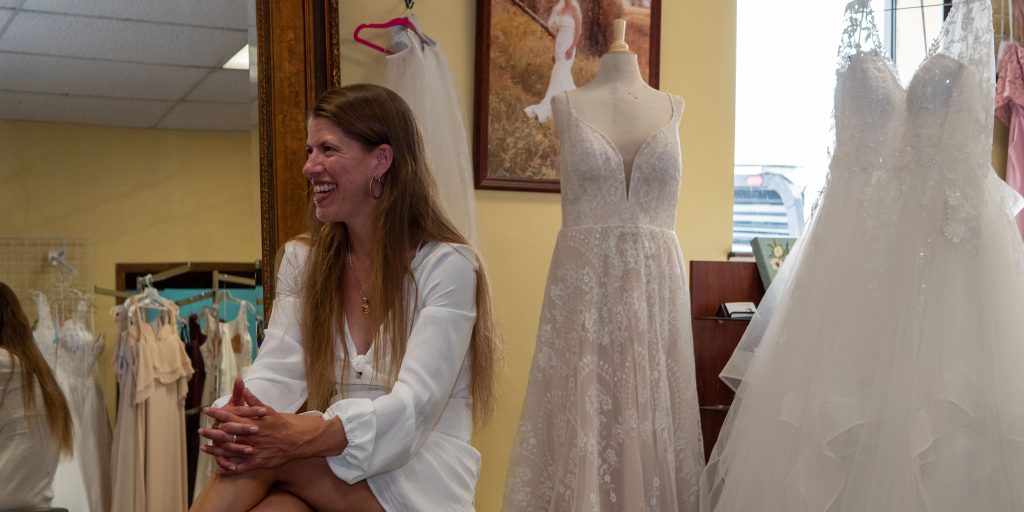 smiling woman with long brown hair wearing white dress, hoop earrings, and necklace sits on a stool in front of a wood-rimmed mirror and bridal gowns on display