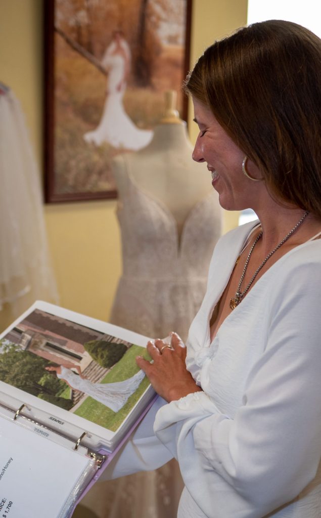 woman with brown hair and silver hoop earrings and necklace smiles as she pages through a pink binder of photos of bridal gowns