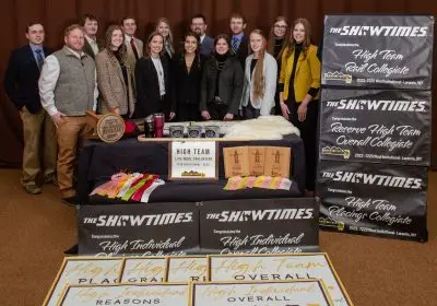 fourteen smiling people wearing formal clothing stand behind a table with wool judging ribbons and plaques. Several banners are arranged in front of the table and on the right side of the group.