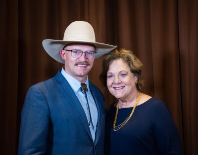 man wearing glasses, light tan cowboy hat and blue jacket with colored shirt stands beside woman wearing navy blue dress and yellow necklace