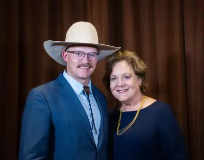 man wearing glasses, light tan cowboy hat and blue jacket with colored shirt stands beside woman wearing navy blue dress and yellow necklace