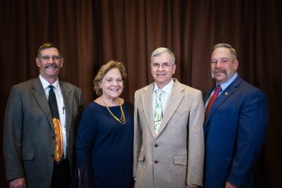 four people stand in a line in front of a brown backdrop. Left to right is a man wearing glasses and a tie with a hotdog, a woman wearing a navy blue dress, a man wearing glasses and a light-colored tie and jacket, and a man with a large mustache wearing a red tie and blue jacket