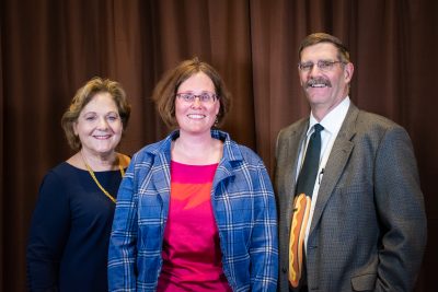 woman in a navy blue dress and yellow necklace poses with woman in a pink shirt and flannel and man wearing glasses and a tie with a hotdog on it