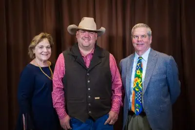 woman wearing navy blue dress and yellow necklace stands beside man wearing cowboy hat and a vest over a red collared shirt. On their right is a man wearing a colorful tie and gray suit jacket.