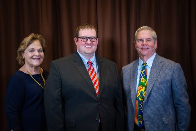 woman wearing navy blue dress and yellow necklace stands beside man wearing suit jacket, glasses, and red tie and another man wearing colorful tie and gray jacket