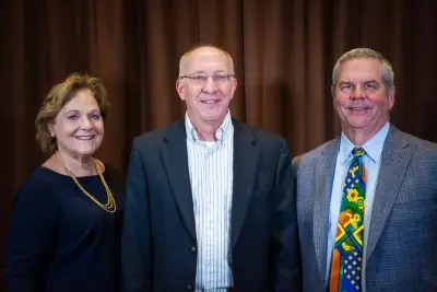 woman in navy blue dress and yellow necklace stands beside man wearing glasses, striped shirt, and navy blue jacket and man wearing gray jacket and colorful tie over a blue shirt