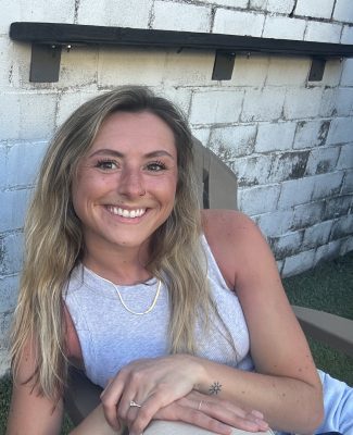 smiling woman with blond hair wearing gray tank top sits in a lawn chair in front of a white cinderblock wall