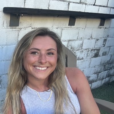 smiling woman with blond hair wearing gray tank top sits in a lawn chair in front of a white cinderblock wall