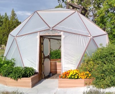 geodesic dome surrounded by green plants and yellow and orange flowers. The door is open and more planters filled with plants can be seen inside.