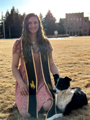 smiling woman wearing red dress and brown and gold graduation stole kneels on brown grass with her hand on a black and white dog