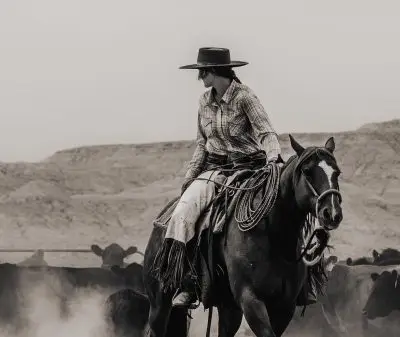 black and white photo of a woman on horseback wearing a cowboy hat, plaid shirt, and chaps looking over her shoulder at cows as dust rises from the ground.
