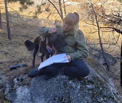 woman wearing an olive green jacket sits on a rock in the woods with an open notebook in her lap and an arm around a black dog wearing a harness.