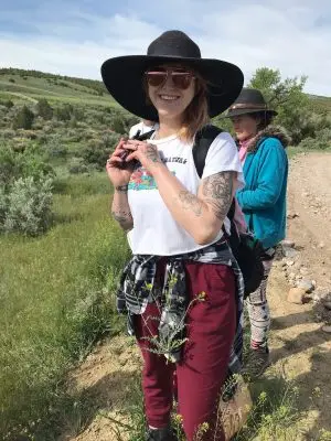 woman wearing black sunhat and aviator sunglasses smiles at the camera on a rocky trail with green hills in the background