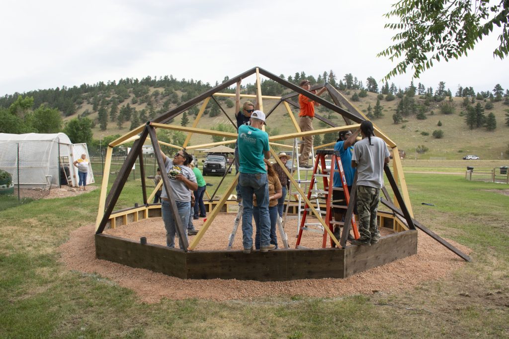 Men and women help hold up pieces of a wooden geodome frame. Three stand on ladders and several others assist from the ground.