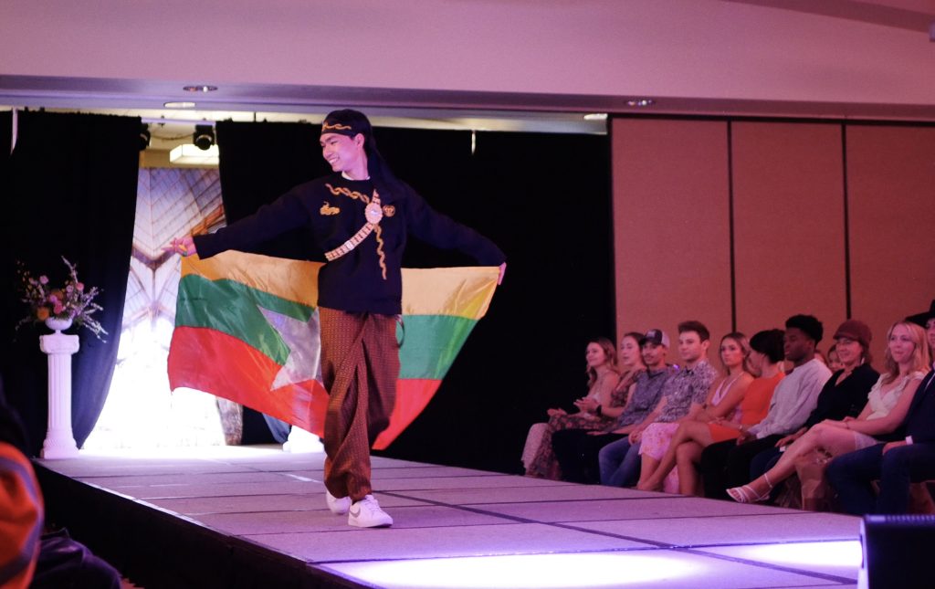 smiling student carrying a Myanmar flag walks down a fashion show runway wearing a black cap and shirt embroidered with gold and baggy pants with a red print.