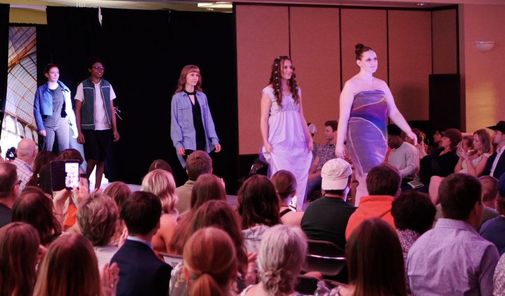 five students walk single file down a fashion show runway surrounded by onlookers