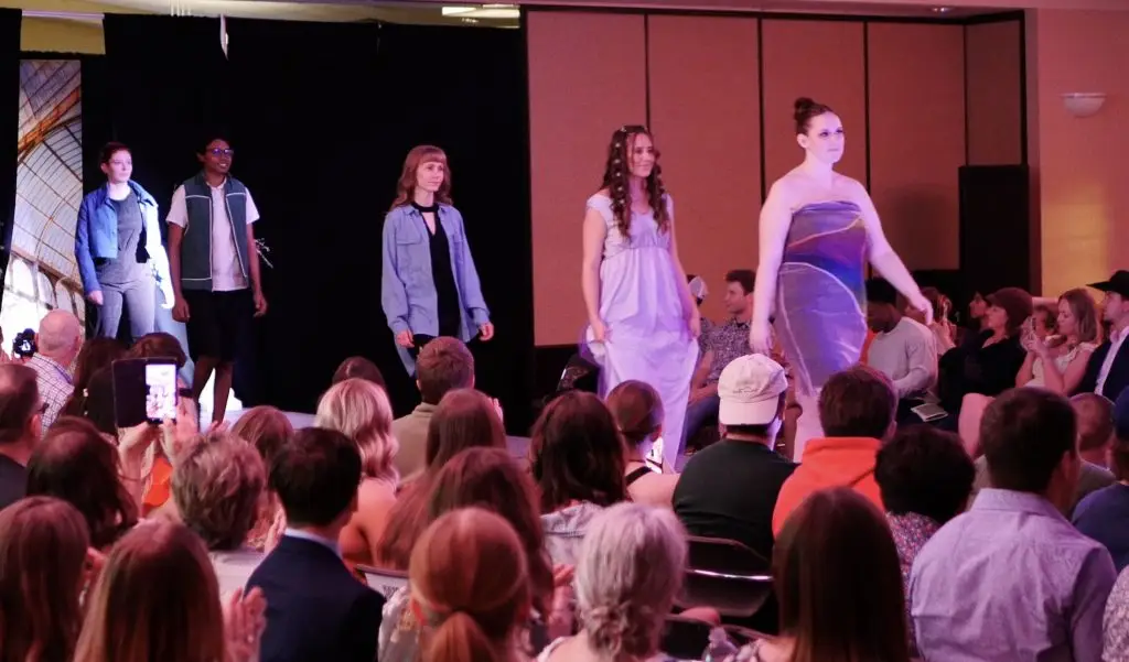 five students walk single file down a fashion show runway surrounded by onlookers