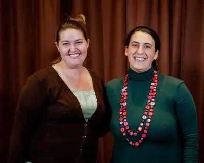smiling woman wearing a brown cardigan and green shirt stands beside smiling woman wearing dark green turtleneck and red and pink necklace.