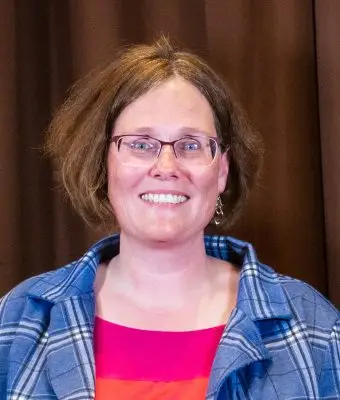 headshot of a smiling woman with short brown hair and glasses wearing a blue flannel over a pink shirt