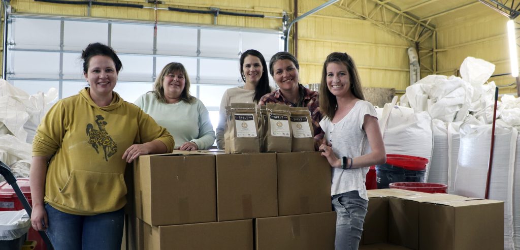 five smiling women stand in front of a closed garage door, clustered around cardboard boxes with brown bags labeled spelt on top. Tall white bags for grain stand to their left.