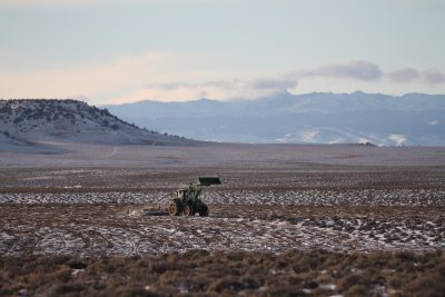 Tree tractor with bucket raised pulls mowing device in a sagebrush field dotted by snow. Wispy clouds rise above snowy peaks in the background.