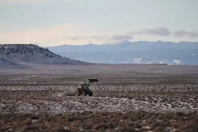 Tree tractor with bucket raised pulls mowing device in a sagebrush field dotted by snow. Wispy clouds rise above snowy peaks in the background.