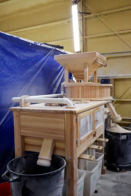 wooden grain mill device with storage bins below and two black garbage cans, one full of spelt flour