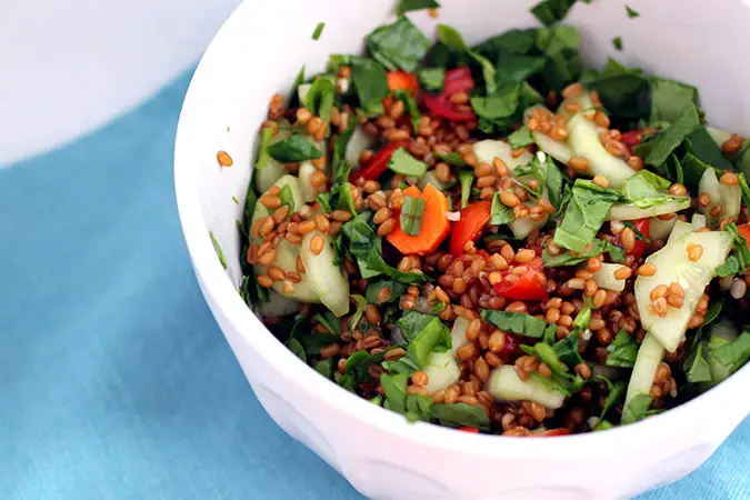 white bowl of salad placed on a light blue cloth. The salad contains greens, cucumbers, tomatoes, carrots, and wheat berries.