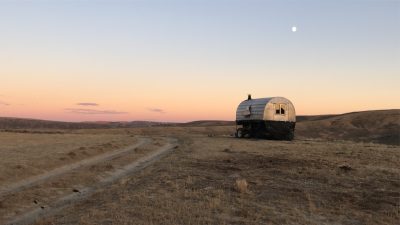 sheepherder's wagon with metal domed top and black bottom sitting off to the side of a dirt road on brown grass at sunset with a moon overhead