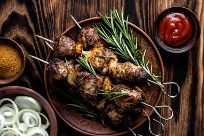 four lamb kebabs adorned with green herbs on a plate on a wooden table with a small bowl of spices, small bowl of raw onion, and small bowl of red dipping sauce