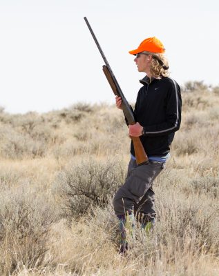 man wearing orange baseball gap, black quarter zip, and gray cargo pants, and hearing protection stands in teh sagebrush holding a hunting rifle.