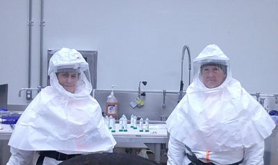 two people wearing personal protective equipment, including white hoods and clear face shields stand in a biosafety level three laboratory space in front of a metal counter containing test tubes and other instrumentation.