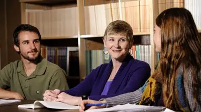 Three people sitting in a library next to each other. A young woman and young man are facing towards each other, smiling. In between them, there is a smiling older woman. They all have notebooks and books in front of them.