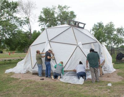 six people work together to secure the opaque plastic skin of a geodesic dome greenhouse to its frame. The dome is located in a gravelly patch surrounded by green grass, picnic tables, and young trees.