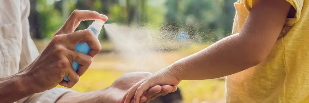close-up of adult spraying light blue bottle of bug spray onto a child's hand and arm.